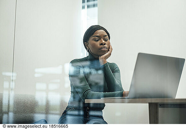 Businesswoman leaning on elbow watching laptop seen through window