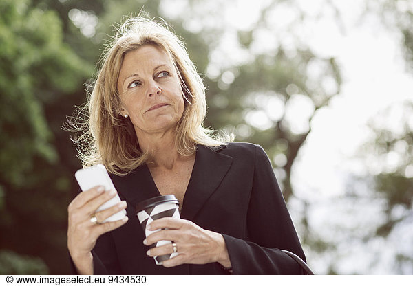 Businesswoman looking away while holding smart phone and disposable coffee cup outdoors