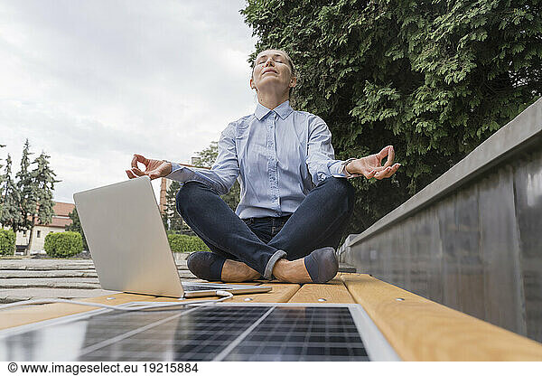 Businesswoman meditating in Lotus position near laptop on bench