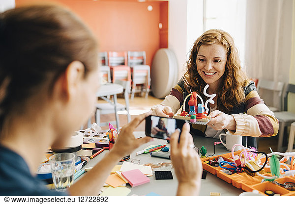 Businesswoman photographing smiling female colleague holding toy at table in creative office