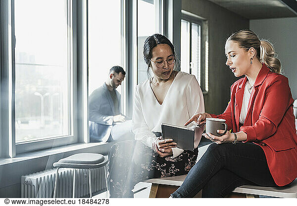 Businesswoman pointing at tablet PC held by colleague sitting on desk in office