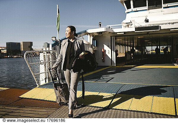 Businesswoman pulling wheeled luggage while disembarking from ferry on sunny day