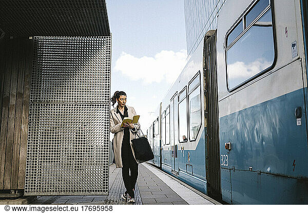 Businesswoman reading book while leaning at metal wall by train at railroad station
