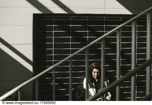 Businesswoman sitting against wall at railroad station on sunny day