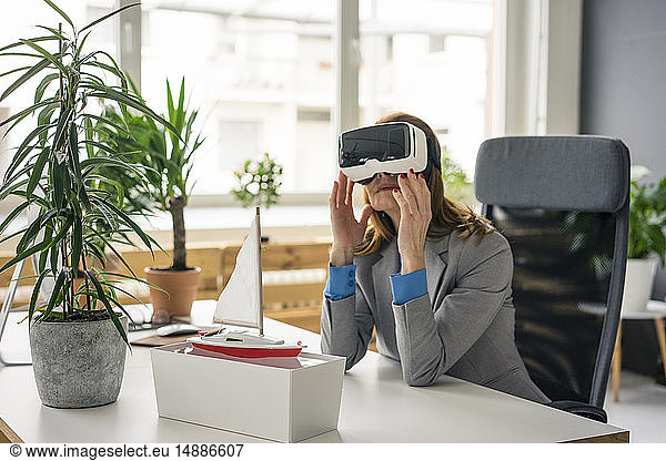Businesswoman sitting at desk with a ship model looking through VR glasses