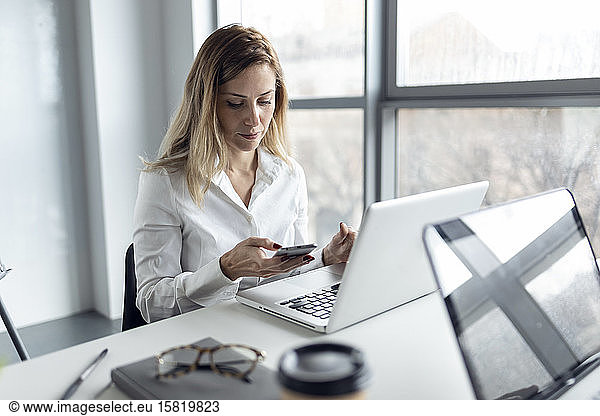 Businesswoman sitting in office working on laptop  using smartphone