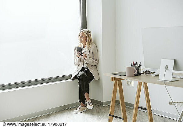 Businesswoman sitting on window sill in office holding mobile phone