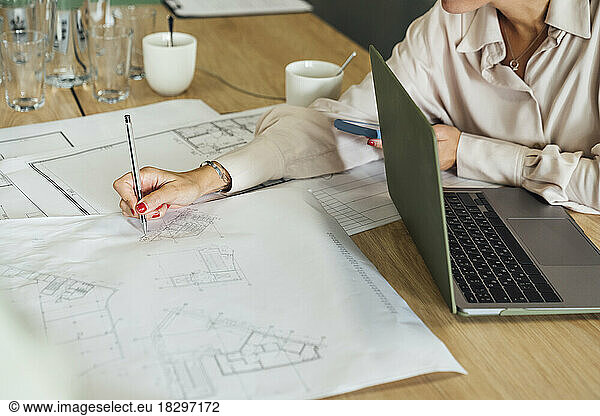 Businesswoman sitting with laptop and working on blueprint at table
