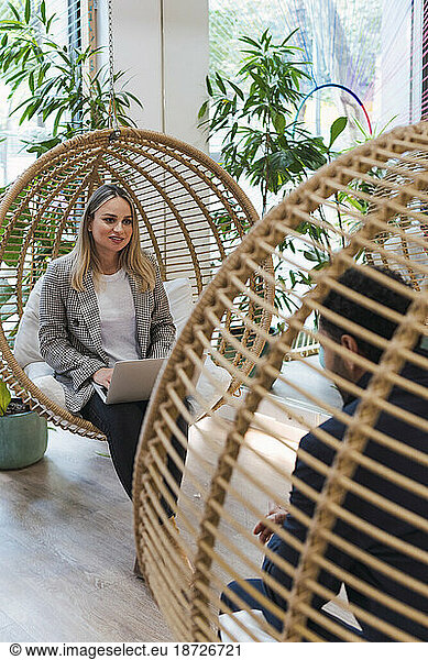 Businesswoman sitting with laptop in hanging chair talking to businessman