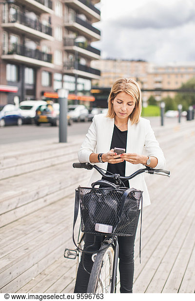 Businesswoman standing with bicycle while using smart phone in city