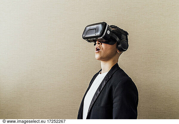 Businesswoman standing with mouth open while using virtual reality headset by wall in office