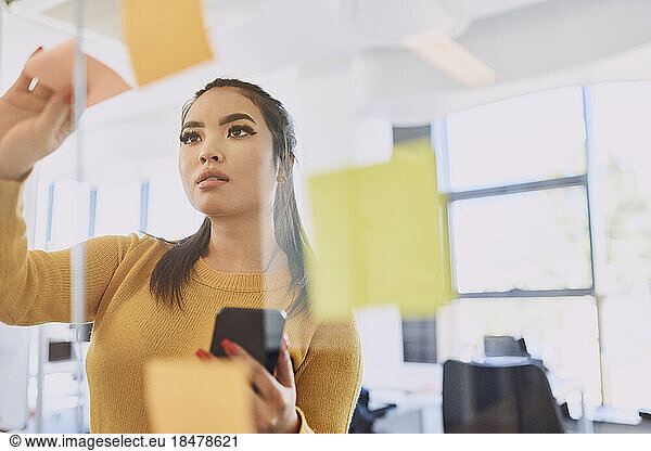 Businesswoman sticking adhesive note on glass wall at workplace