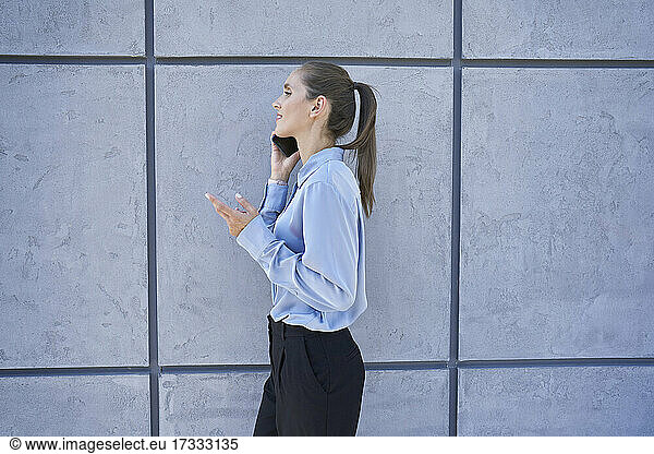 Businesswoman talking on mobile phone while walking by wall