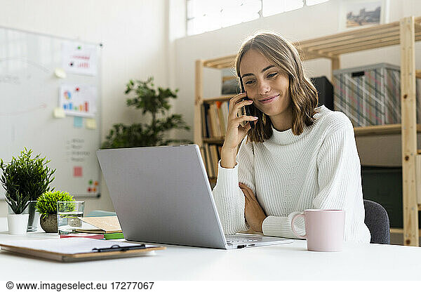 Businesswoman talking on mobile phone while working on laptop at office