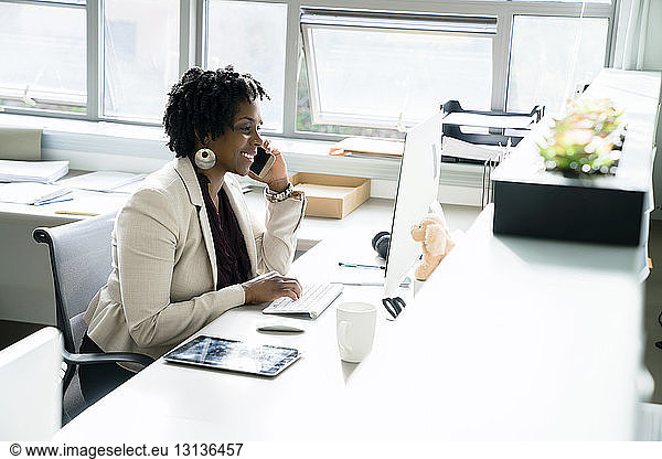 Businesswoman talking on smart phone while using desktop computer in office