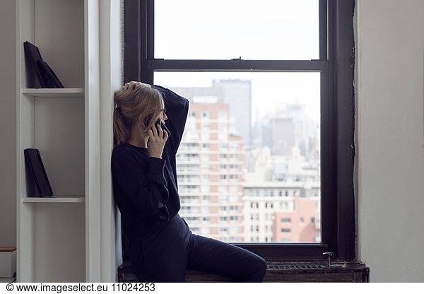 Businesswoman talking through smart phone while sitting on window sill at city