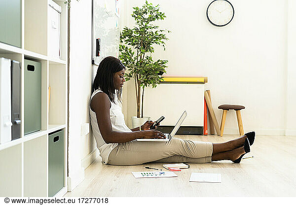 Businesswoman using laptop and mobile phone together while sitting on floor at office
