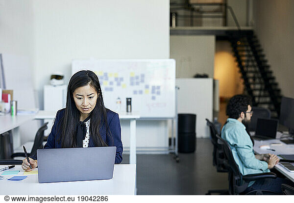 Businesswoman using laptop computer while male colleague working at desk in office