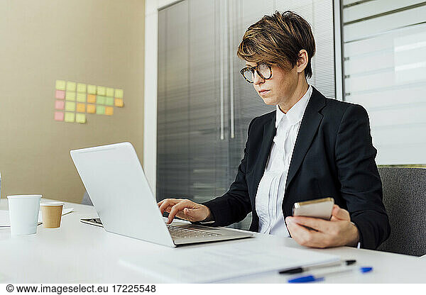 Businesswoman using laptop while holding mobile phone at desk in office
