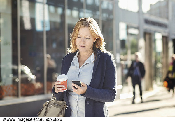 Businesswoman using mobile phone and holding disposable coffee cup while walking on sidewalk