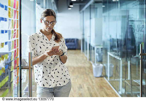 Businesswoman using mobile phone while leaning on glass door at corridor