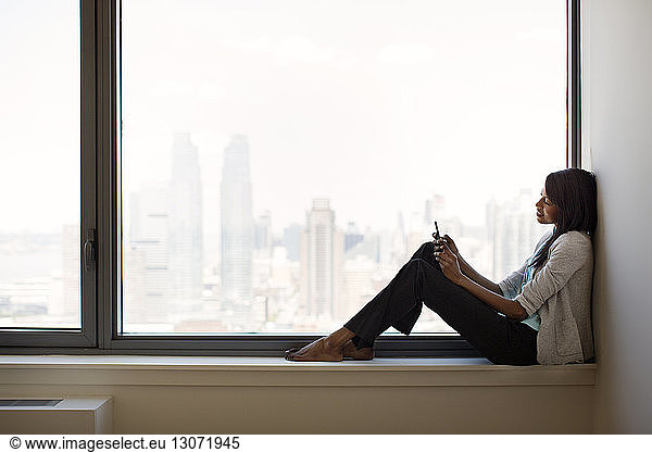 Businesswoman using mobile phone while sitting on window sill in office
