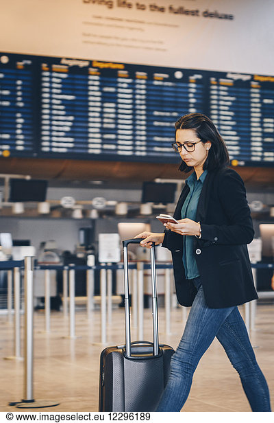 Businesswoman using mobile phone while walking with luggage in airport terminal