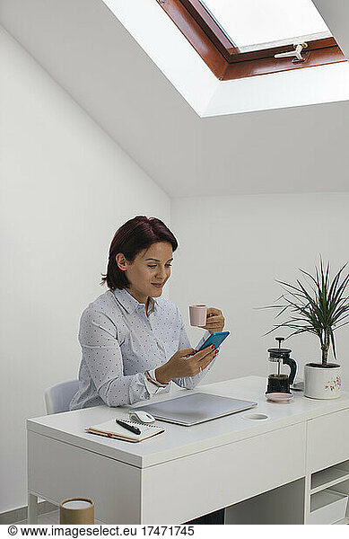 Businesswoman using smart phone while having coffee at desk in home office
