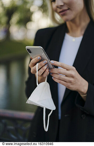 Businesswoman using smart phone while holding protective mask in city