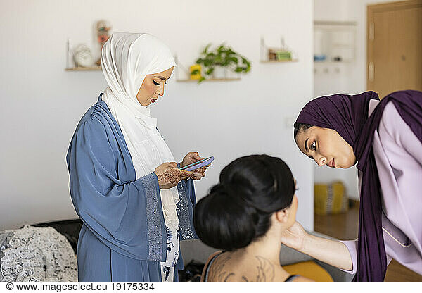 Businesswoman using smart phone with make-up artist preparing bride for wedding at home