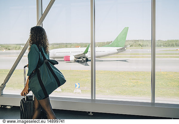 Businesswoman walking with luggage by window in corridor at airport