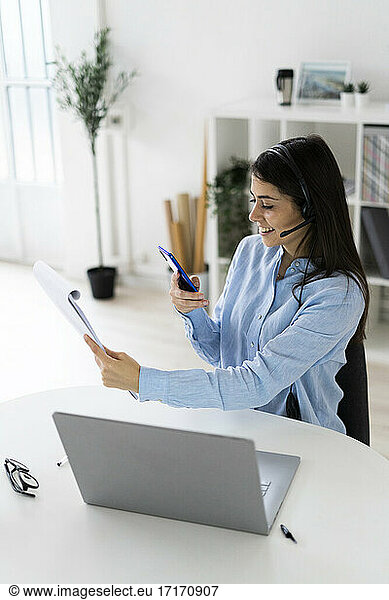 Businesswoman wearing headset taking photo of note pad through mobile phone while sitting by desk at office