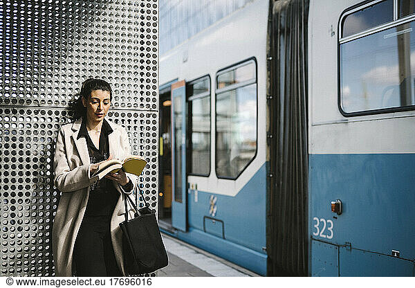 Businesswoman wearing long coat reading book while waiting at railroad station