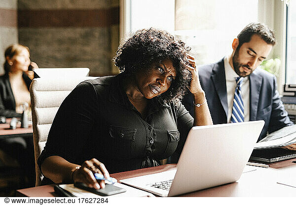 Businesswoman with hand in hair using laptop by businessman working at office