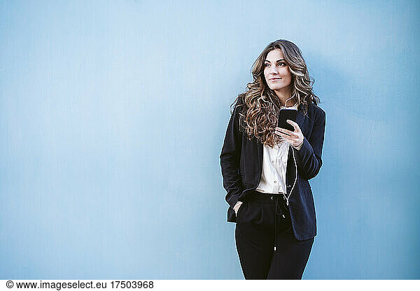 Businesswoman with hand in pocket holding smart phone in front of blue wall