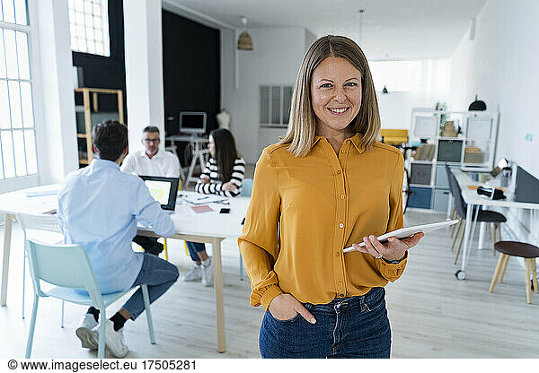Businesswoman with hand in pocket holding tablet PC with colleagues sitting at office