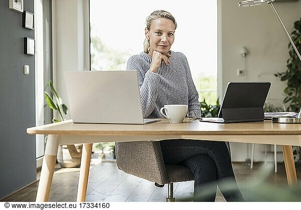 Businesswoman with hand on chin sitting by desk at home office