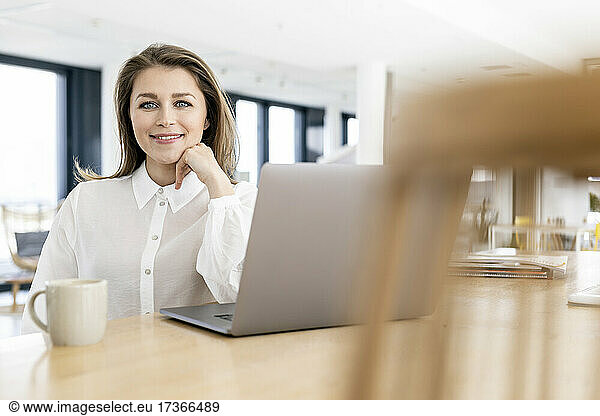 Businesswoman with hand on chin sitting by laptop at office