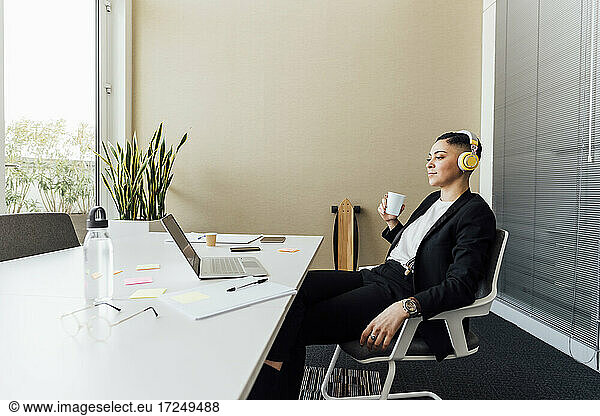 Businesswoman with headphones and coffee cup looking at laptop in office