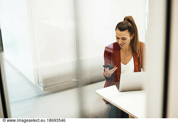 Businesswoman with laptop using smart phone sitting at desk in office