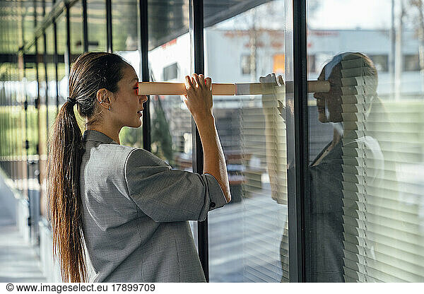 Businesswoman with long ponytail looking at reflection through rolled up paper on glass wall