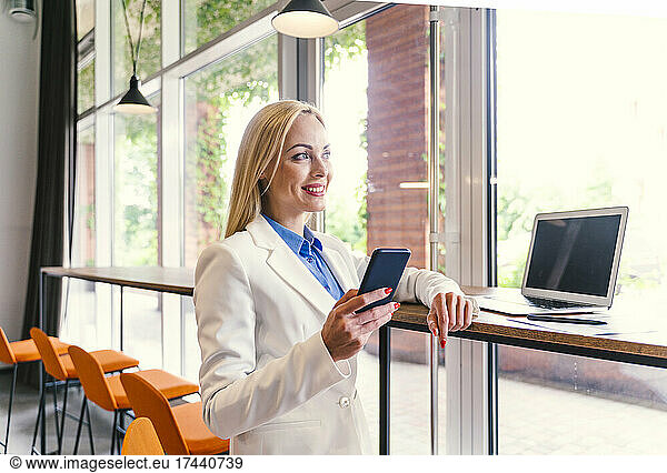 Businesswoman with mobile phone and laptop standing at desk in office