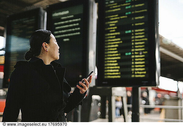 Businesswoman with smart phone looking at departure board
