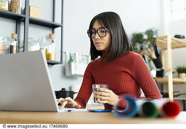 Businesswoman working on laptop holding water glass at home office