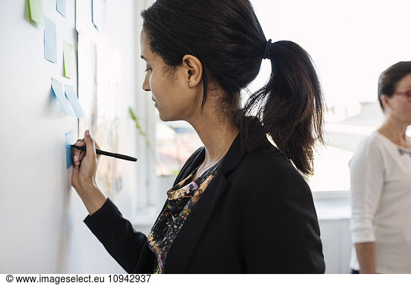 Businesswoman writing in adhesive note on wall at board room