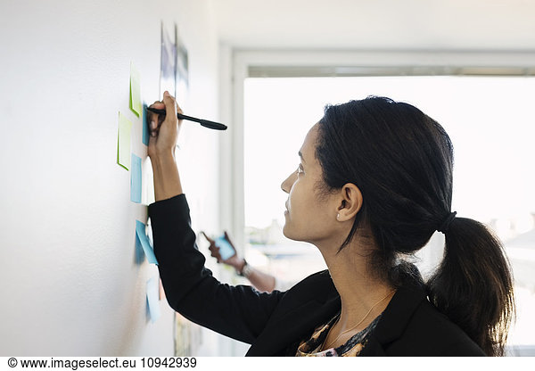 Businesswoman writing in adhesive note on wall in office