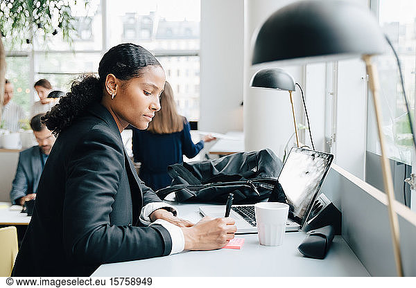 Businesswoman writing notes while working on laptop at desk in coworking space