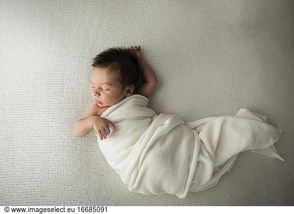 Candid Newborn Baby With Long Hair Sleeps in White Swaddle