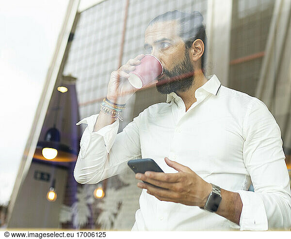 Candid portrait of bearded businessman drinking coffee and using phone inside cafe