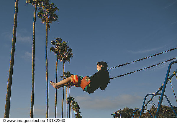 Carefree boy swinging at playground against sky and coconut palm trees